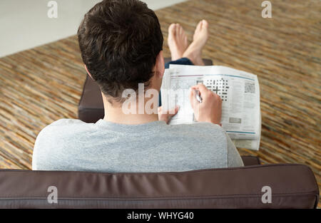 Rear view of a young man sitting on sofa and doing crossword puzzle in newspaper Stock Photo