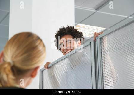 Smiling male office worker peering over cubicle wall to greet blond coworker in office Stock Photo