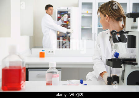 Scientist using microscope with male colleague looking in refrigerator in laboratory Stock Photo