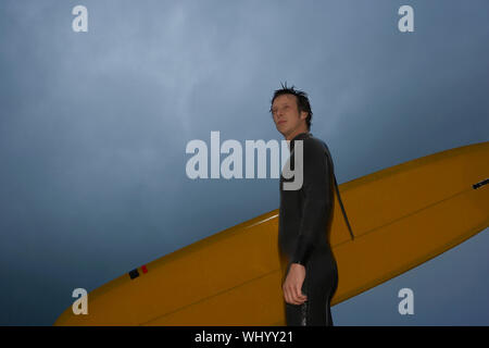 Low angle view of confident young man with surfboard looking away against cloudy sky at beach Stock Photo