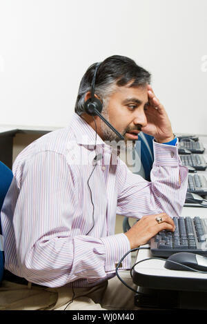 Side view of a tired customer service operator communicating on headset while typing on keyboard Stock Photo