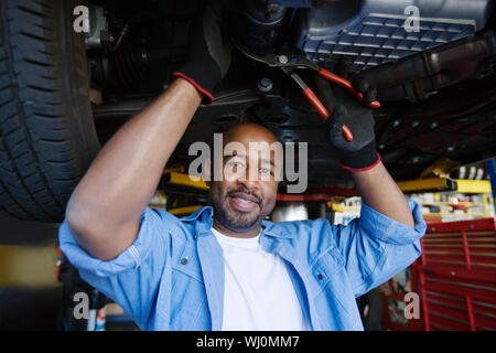 Portrait of a happy African American male mechanic repairing a lifted car In Garage Stock Photo