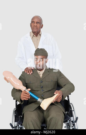 Senior doctor standing behind military officer holding artificial limb as he sits in wheelchair over gray background Stock Photo