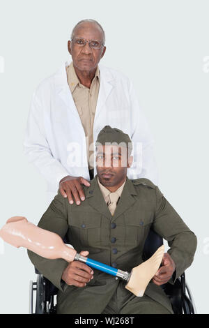 Portrait of senior doctor standing with military officer holding artificial limb as he sits in wheelchair over gray background Stock Photo