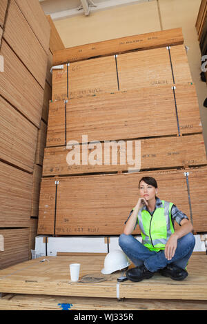 Pensive female industrial worker sitting cross-legged on wooden planks Stock Photo