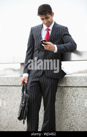 Young Indian businessman texting through cell phone while holding laptop bag at parapet Stock Photo