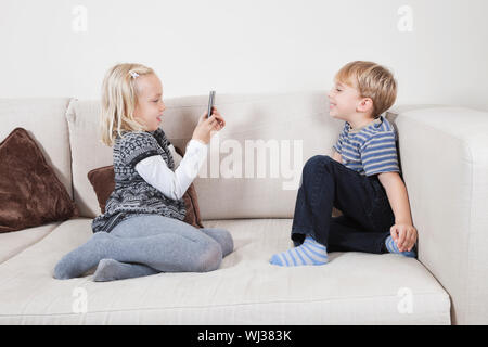 Young girl photographing brother through cell phone on sofa Stock Photo