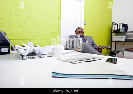 Indian businessman asleep at his desk clutching ukulele Stock Photo