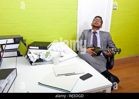 Indian businessman asleep at his desk clutching ukulele Stock Photo