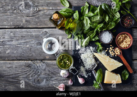 homemade Basil pesto sauce in a glas jar with fresh basil leaves, parmesan cheese, pine nuts, garlic, peppercorns, bottle of olive oil on a rustic woo Stock Photo