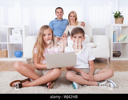 Children On The Carpet Using Tablet And Laptop With Parents Behind Them At Home Stock Photo