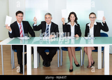 Group of four stylish professional judges seated at a long table holding up blank cards for their scores Stock Photo