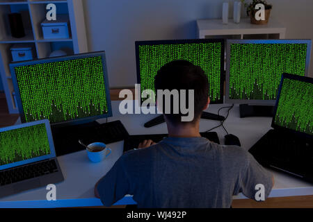 Rear high angle view of a male hacker sitting at a desk surrounded by computer monitors streaming data as he steals information Stock Photo