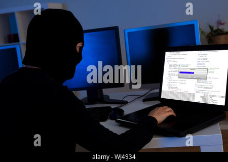 Masked hacker wearing a balaclava sitting at a desk downloading private information off a computer Stock Photo