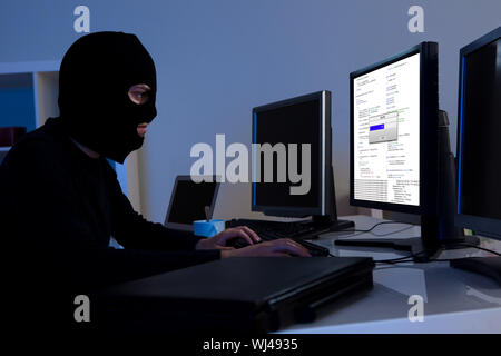 Masked hacker wearing a balaclava sitting at a desk downloading private information off a computer Stock Photo