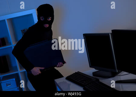 Man dressed in black and wearing a balaclava is stealing a laptop from an office Stock Photo