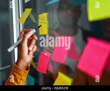 Close-up of businesswoman hand writing on yellow sticky notes stuck over glass board in creative office Stock Photo
