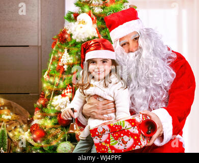 Portrait of little girl receive gift box from Santa Claus, sitting near decorated Christmas tree, happy childhood, Christmastime children's party Stock Photo