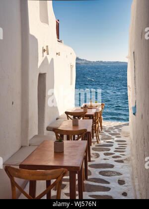 Brown tables and chairs of street restaurant placed in narrow passage between building near rippling sea on Mykonos Island in Greece Stock Photo