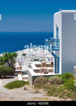 Green shrubs growing in yard of building with white walls and blue balcony against coastal city and blue sea on Mykonos Island in Greece Stock Photo