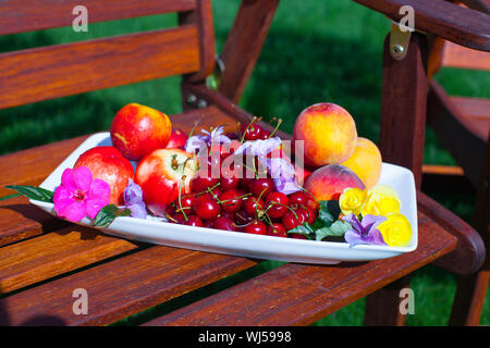 Plate with fresh fruits and flowers on wooden chairs in the garden Stock Photo