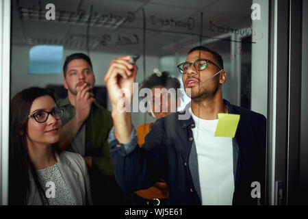Pensive diverse group of young people looking at businessman writing business future plans on glass during office meeting Stock Photo