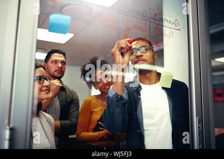 Multi ethnic happy creative business team looking at young man writing ideas on glass board with red marker at office Stock Photo