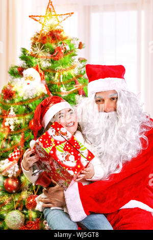 Little happy girl receive gift from Santa Claus, sitting near beautiful decorated Christmas tree, enjoying Xmas eve at home Stock Photo