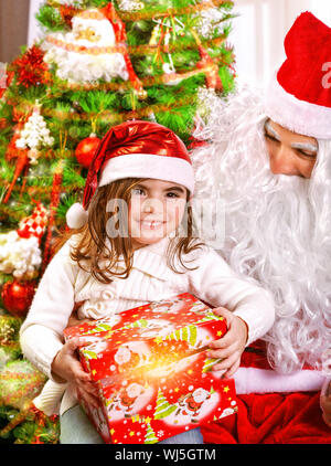 Little happy girl receive gift from Santa Claus, sitting near beautiful decorated Christmas tree, enjoying Xmas eve at home Stock Photo