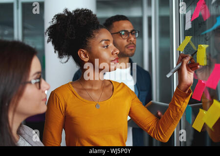 Young creative businesswoman standing with her colleagues writing new ideas on sticky notes over glass wall Stock Photo