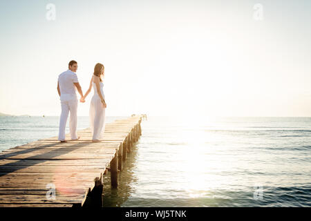 Rear View Of Couple Holding Hands While Walking On Wooden Jetty Over Sea Against Sky During Sunny Day