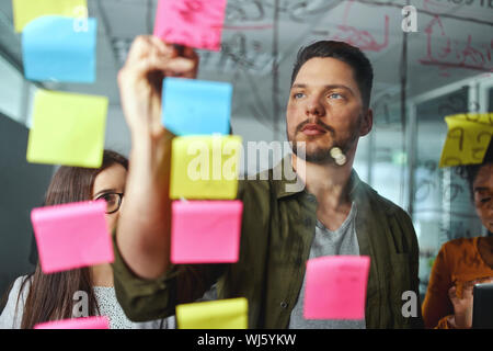 Concentrated male executive discussing business ideas with colleagues by writing on sticky notes pasted over the glass board Stock Photo