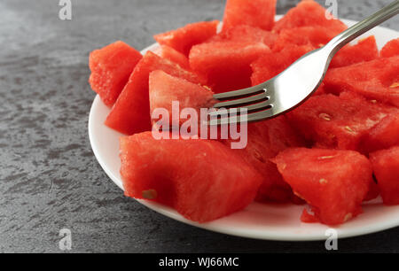 Side view of a plate of watermelon chunks with a fork spearing a piece on a gray mottled table illuminated with natural lighting. Stock Photo