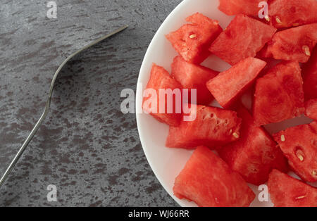 Top close view of a plate of watermelon chunks with a fork to the side on a gray mottled table illuminated with natural lighting. Stock Photo