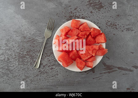 Top view of a plate of watermelon chunks with a fork to the side on a gray mottled table illuminated with natural lighting. Stock Photo