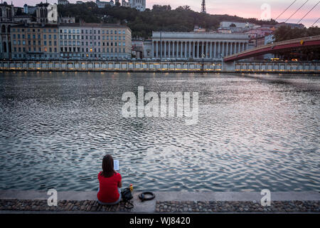 LYON, FRANCE - JULY 17, 2019: Young woman reading a book & relaxing on the riverbank of Saone river in Lyon, at dusk, with Colline de Fourviere hill b Stock Photo