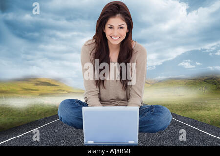 Composite image of a woman smiling as she looks forward in front of her laptop with crossed legs. Stock Photo