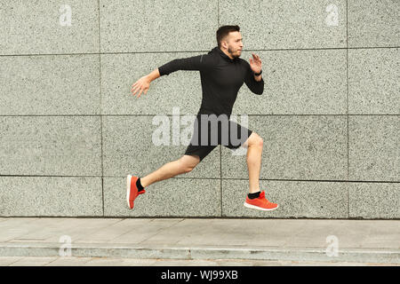Young man jogging in city against grey urban wall, copy space, side view Stock Photo