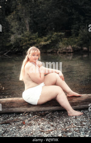 Young curvy woman sits scantily clad on a lake on a tree trunk Stock Photo