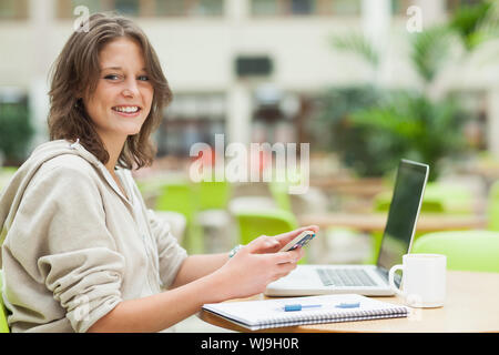 Portrait of a female student using cellphone and laptop at cafeteria table Stock Photo