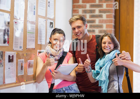 Three smiling students standing next to notice board showing thumbs up in school Stock Photo
