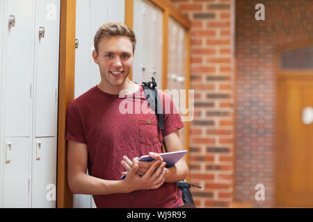 Handsome smiling student leaning against lockers using tablet in school Stock Photo