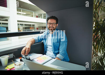 Smiling creative professional going over notes and working on a laptop while sitting alone in an office pod Stock Photo