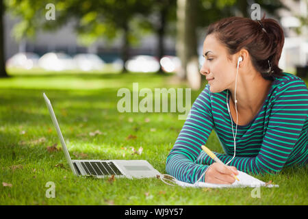 Attractive casual student lying on grass taking notes on campus at college Stock Photo