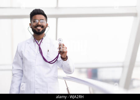 Portrait of a smiling Asian Indian male medical doctor in uniform, hospital building at background. Stock Photo
