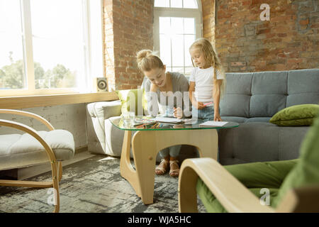 Female caucasian teacher and little girl, or mom and daughter. Homeschooling. Sitting on the sofa and drawing with paints, writing, talking and having fun. Education, school, studying concept. Stock Photo