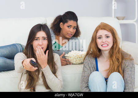 Portrait of scared young female friends with remote control and popcorn bowl on sofa at home Stock Photo