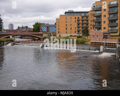 Red bridge Orange boom weir side flow on the river Aire in Leeds with modern flats Leeds Yorkshire England Stock Photo