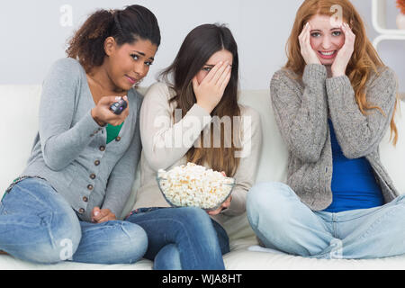 Portrait of scared young female friends with remote control and popcorn bowl on sofa at home Stock Photo