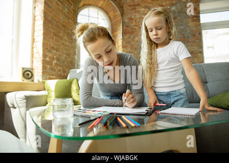 Female caucasian teacher and little girl, or mom and daughter. Homeschooling. Sitting on the sofa and drawing with paints, writing, talking and having fun. Education, school, studying concept. Stock Photo